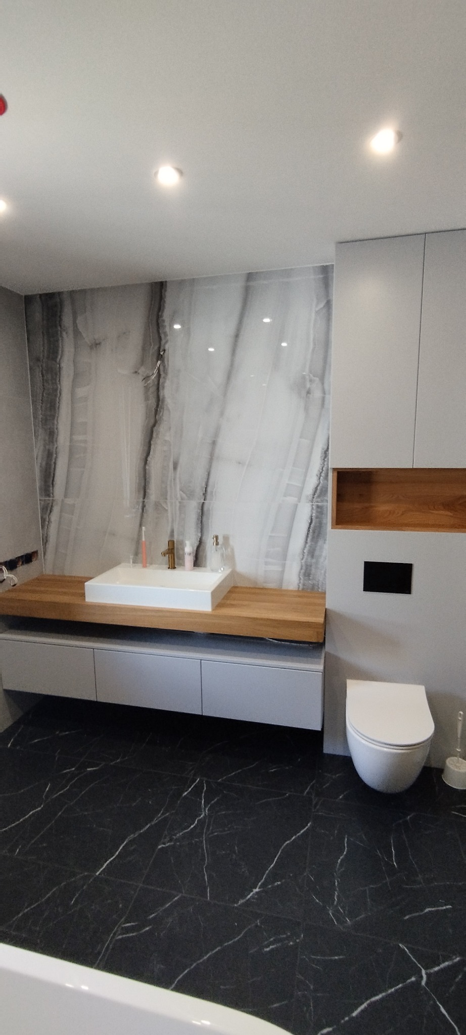 Nowoczesna łazienka z marmurową ścianą, drewnianym blatem pod umywalką i czarną podłogą z białym wzorem. Minimalistyczny design, oświetlenie punktowe.