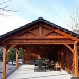 Drewniana altana z ciemnym dachem, widok z zewnątrz, z widocznym stołem i krzesłami wewnątrz, otoczona zielenią i drzewami.