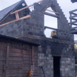 Surowy stan budynku w trakcie wznoszenia ścian z bloczków betonowych, widoczne drewniane elementy konstrukcyjne dachu i otwory okienne, z boku drewniana konstrukcja i rower.