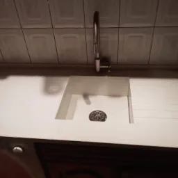 Jasny blat z konglomeratu kwarcowego z wbudowanym zlewozmywakiem, bateria chromowana, tło z płytek ceramicznych.