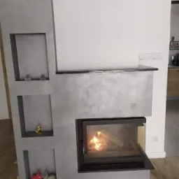 Nowoczesny kominek z szarego betonu architektonicznego, z wnękami na dekoracje i paleniskiem z czarną ramą, płomienie wewnątrz, drewniana podłoga.