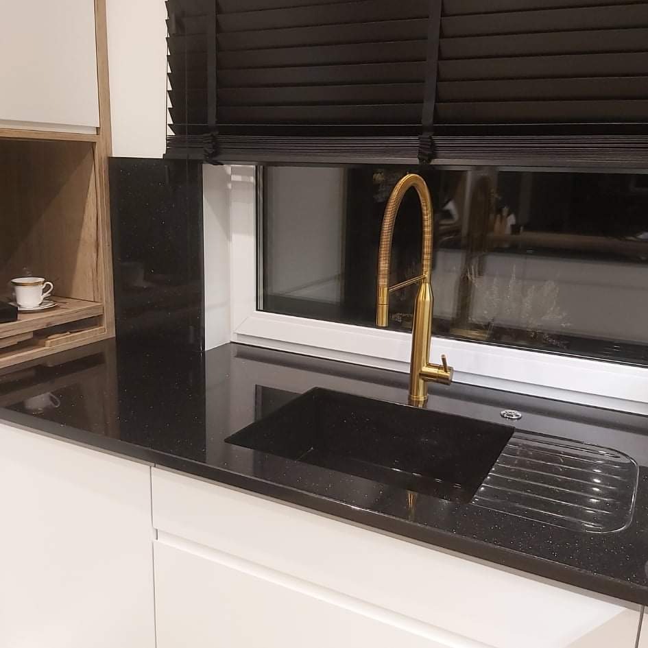 Czarny, błyszczący blat kuchenny z brokatem, złota bateria przy czarnym zlewie, białe szafki i czarne żaluzje w oknie.