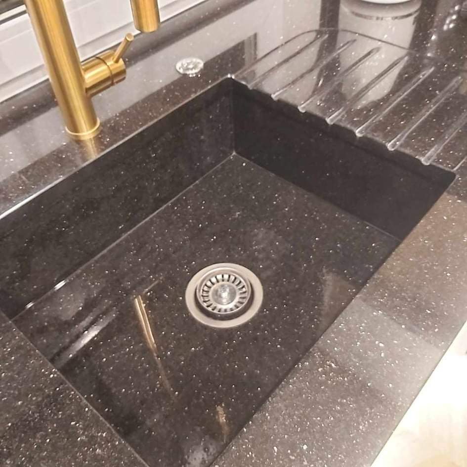 Czarny, brokatowy zlew kuchenny z baterią w kolorze złotym i ociekaczem. Widoczny metalowy odpływ.