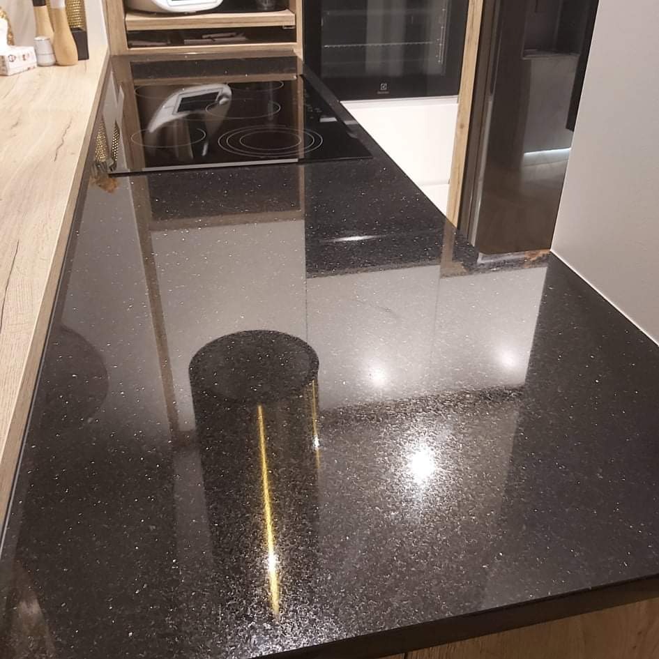 Błyszczący, czarny blat kuchenny z drobnymi, srebrnymi inkluzjami, widoczna płyta indukcyjna i fragment zabudowy kuchennej z jasnego drewna.