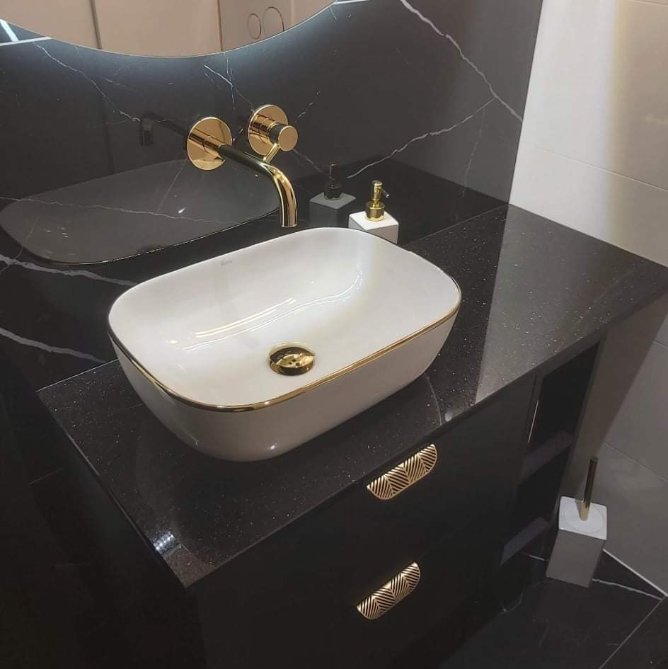 Elegancka łazienka: biała, owalna umywalka z złotym rantem na czarnym blacie z drobinkami, złota bateria ścienna, czarne marmurowe ściany z białymi żyłami, złote uchwyty szafki.
