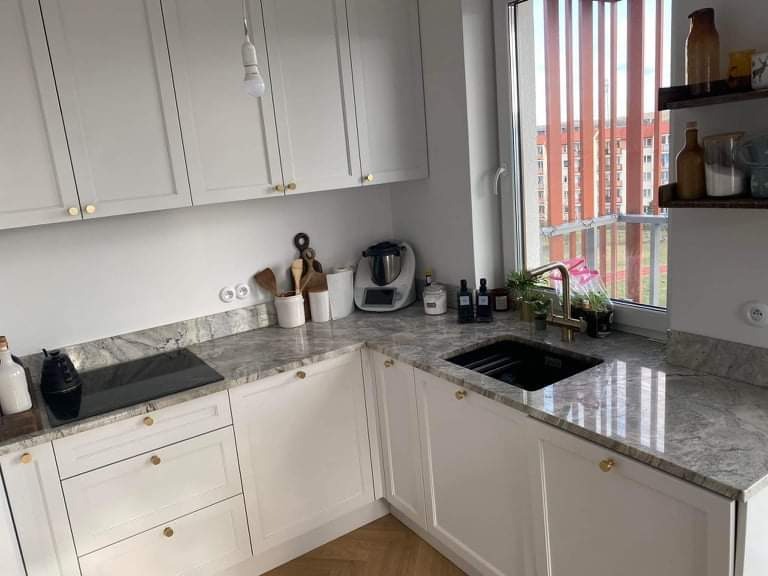 Jasna kuchnia z blatem kamiennym o szarym wzorze, złotymi uchwytami i widokiem na budynek przez okno.