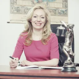 Uśmiechnięta blondynka w różowej bluzce siedzi za biurkiem z posągiem Temidy i otwartym notesem, trzymając długopis w dłoni.