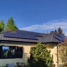 Dom jednorodzinny z zamontowanymi panelami słonecznymi na ciemnym dachu, widok z poziomu ogrodu, okno z żaluzjami odbija zieleń, na pierwszym planie krzewy i kwitnąca magnolia.