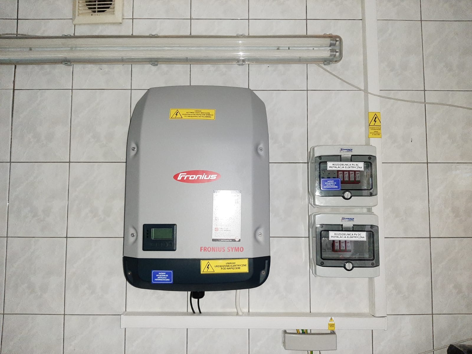 Szary inwerter Fronius Symo zamontowany na ścianie z białych płytek, obok dwie rozdzielnice elektryczne PV AC i PV DC marki Schrack, widoczna lampa jarzeniowa i żółte naklejki ostrzegawcze.