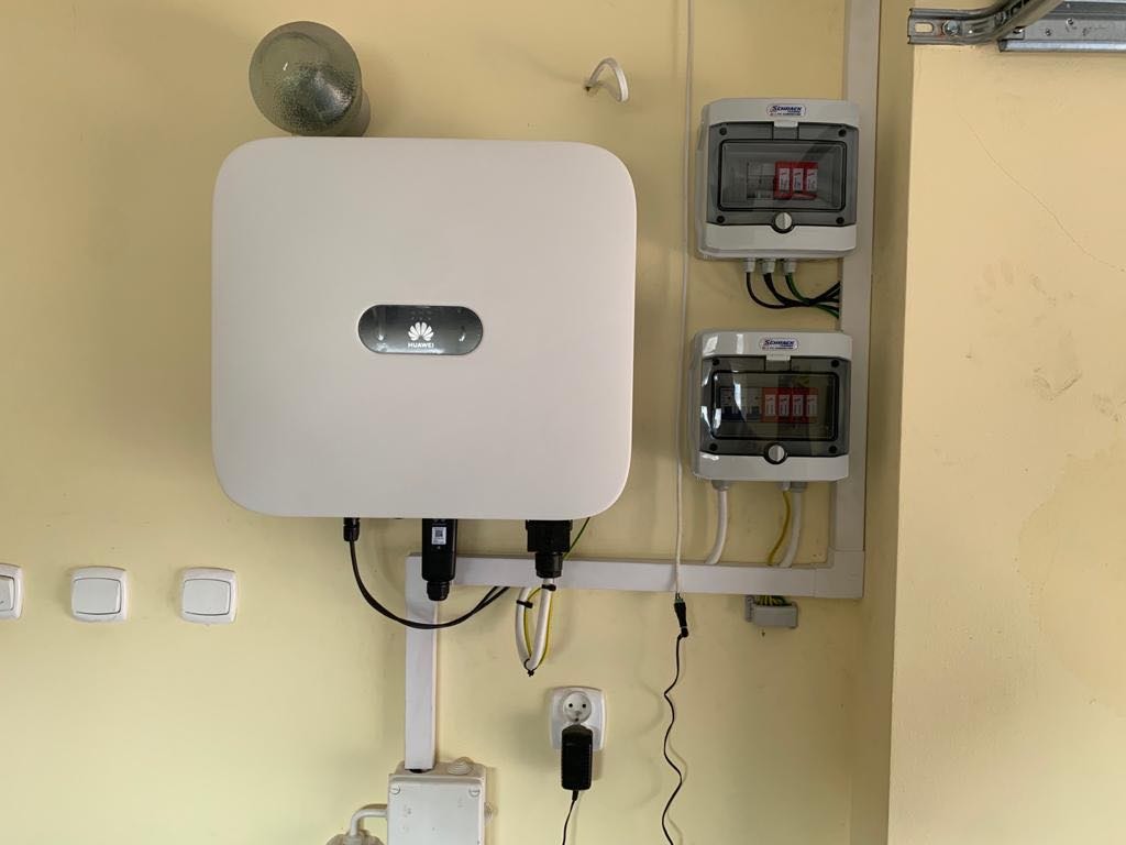 Biały inwerter Huawei zamontowany na żółtej ścianie obok dwóch skrzynek bezpiecznikowych z przezroczystymi pokrywami.