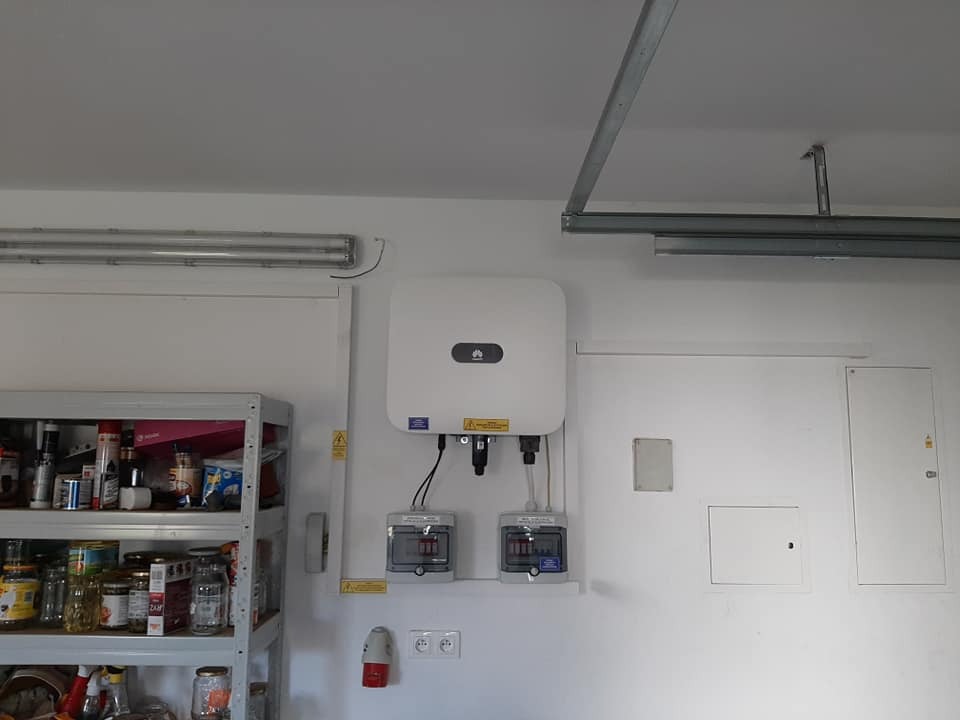 Biały falownik firmy Huawei zamontowany na ścianie w garażu obok metalowego regału z przetworami i skrzynkami elektrycznymi.