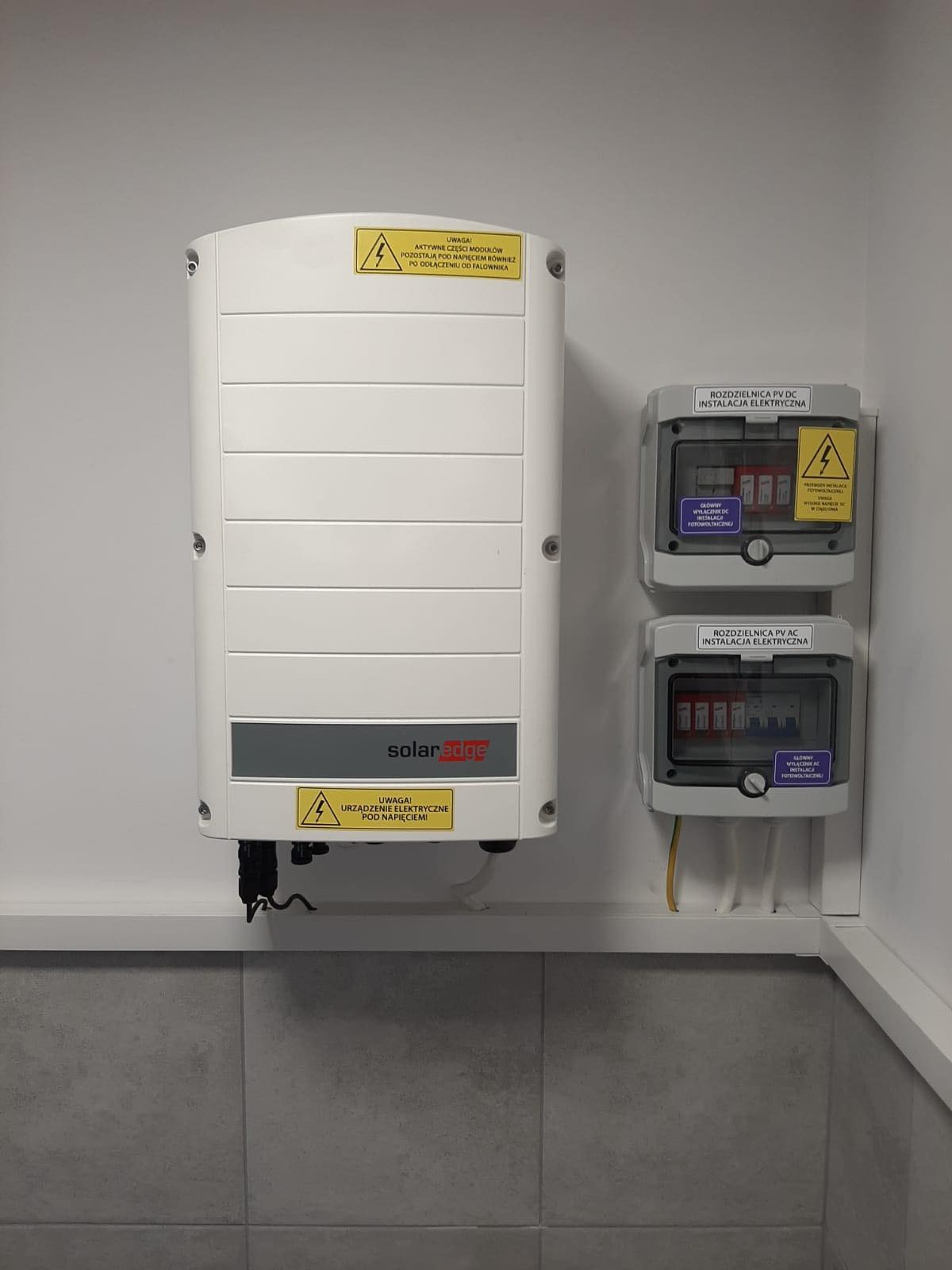 Biały falownik SolarEdge zamontowany na ścianie obok dwóch szarych rozdzielnic PV DC i PV AC instalacji elektrycznej, z żółtymi przewodami w listwie przypodłogowej i szarą płytką ceramiczną w tle.