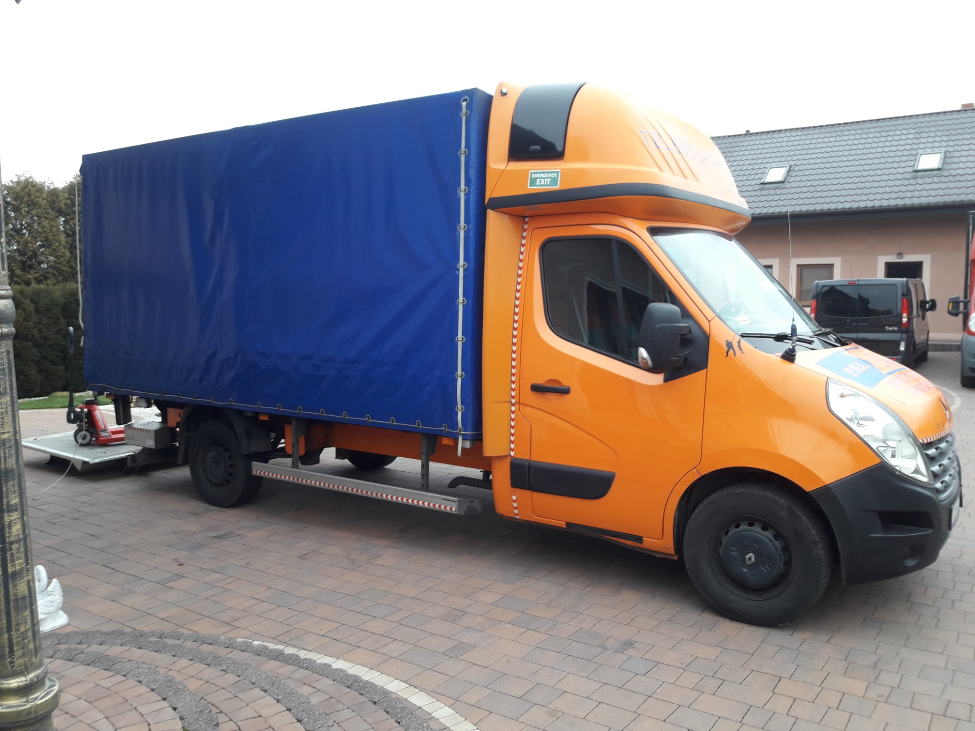 Pomarańczowy samochód dostawczy z niebieską plandeką, windą załadowczą i wózkiem paletowym na platformie, zaparkowany na kostce brukowej. Na kabinie naklejka 'EMERGENCY EXIT'.