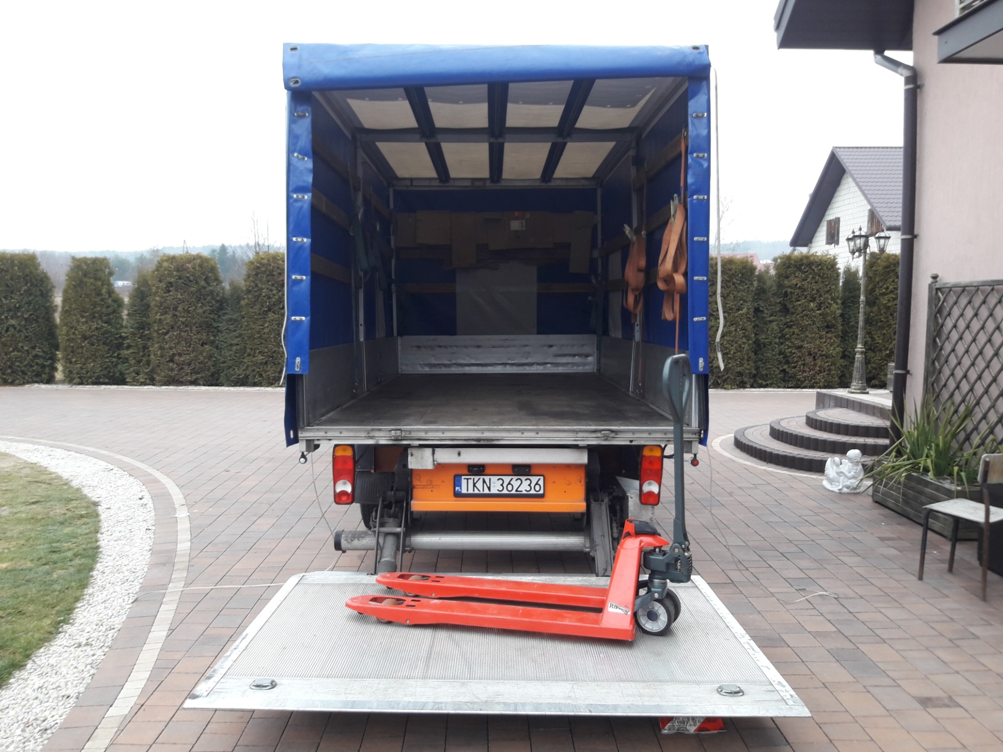 Otwarta pusta paka samochodu dostawczego z windą załadowczą i wózkiem paletowym na platformie, widok z tyłu na tle posesji z tujami i rzeźbą aniołków.