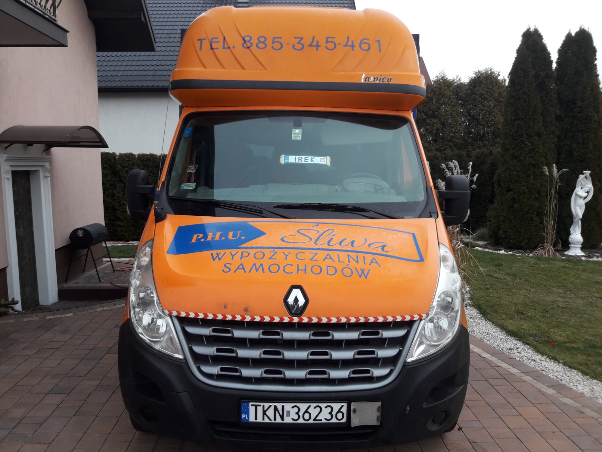 Pomarańczowy samochód dostawczy Renault Master z zabudową transportową, na którym widnieje napis 'WYPOŻYCZALNIA SAMOCHODÓW' i numer telefonu, zaparkowany na brukowanym podjeździe przed domem...