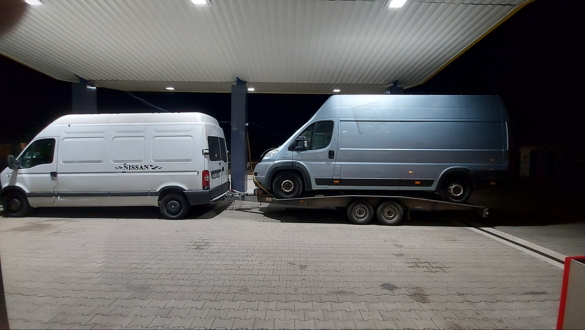 Biały van Nissan ciągnie na lawecie szarego dostawczaka pod zadaszeniem stacji benzynowej.