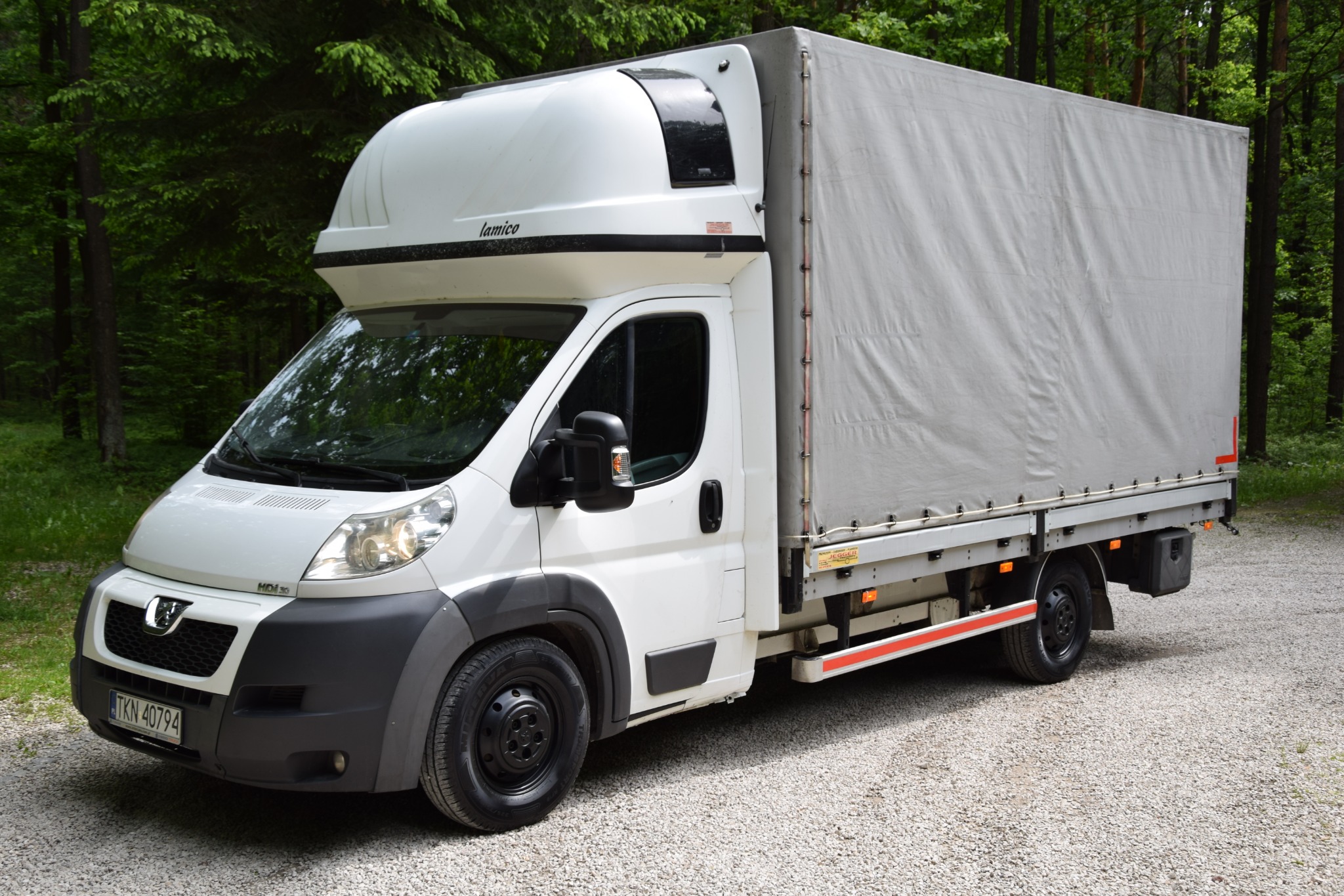 Biały samochód dostawczy z plandeką marki Lamico, zaparkowany na żwirowej drodze na tle zielonego lasu, widoczne światła i tablica rejestracyjna.