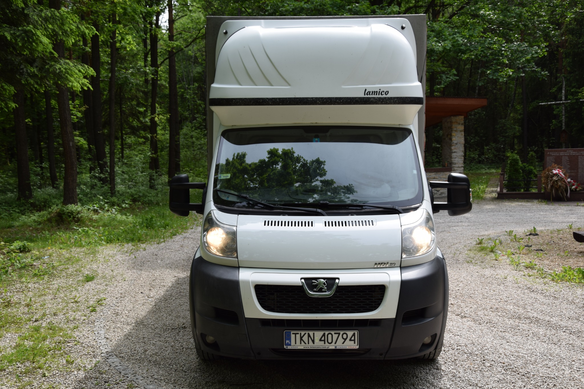 Biały samochód dostawczy marki Peugeot z zabudową Lamico, zaparkowany na żwirowej drodze w pobliżu lasu i pomnika, widok z przodu z włączonymi światłami.