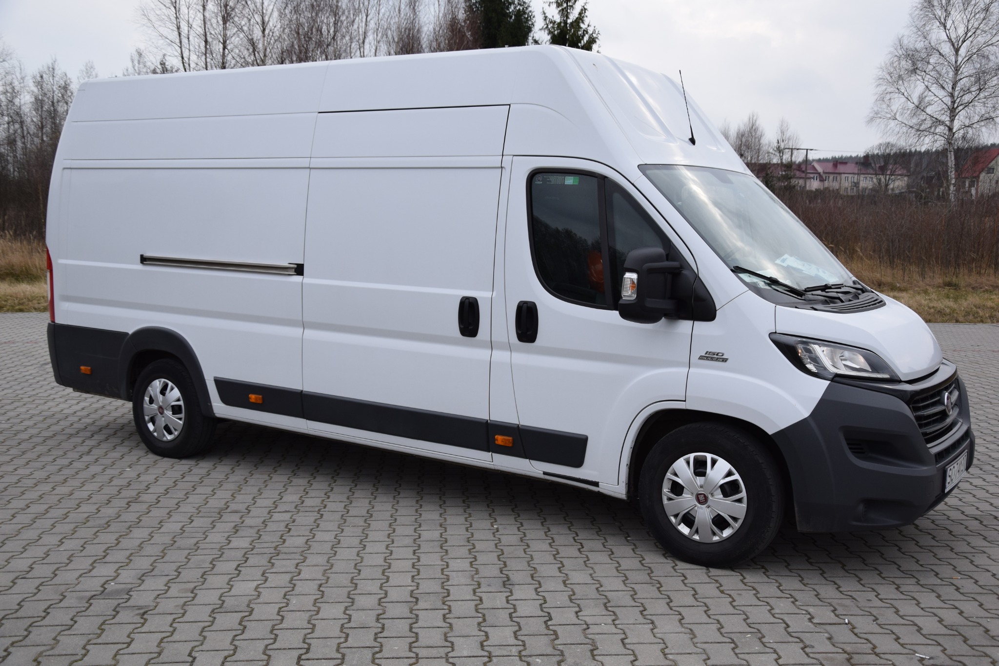 Biały, dostawczy Fiat Ducato z wysokim dachem zaparkowany na szarej kostce brukowej, widok z boku, na przedniej szybie widoczne dokumenty.