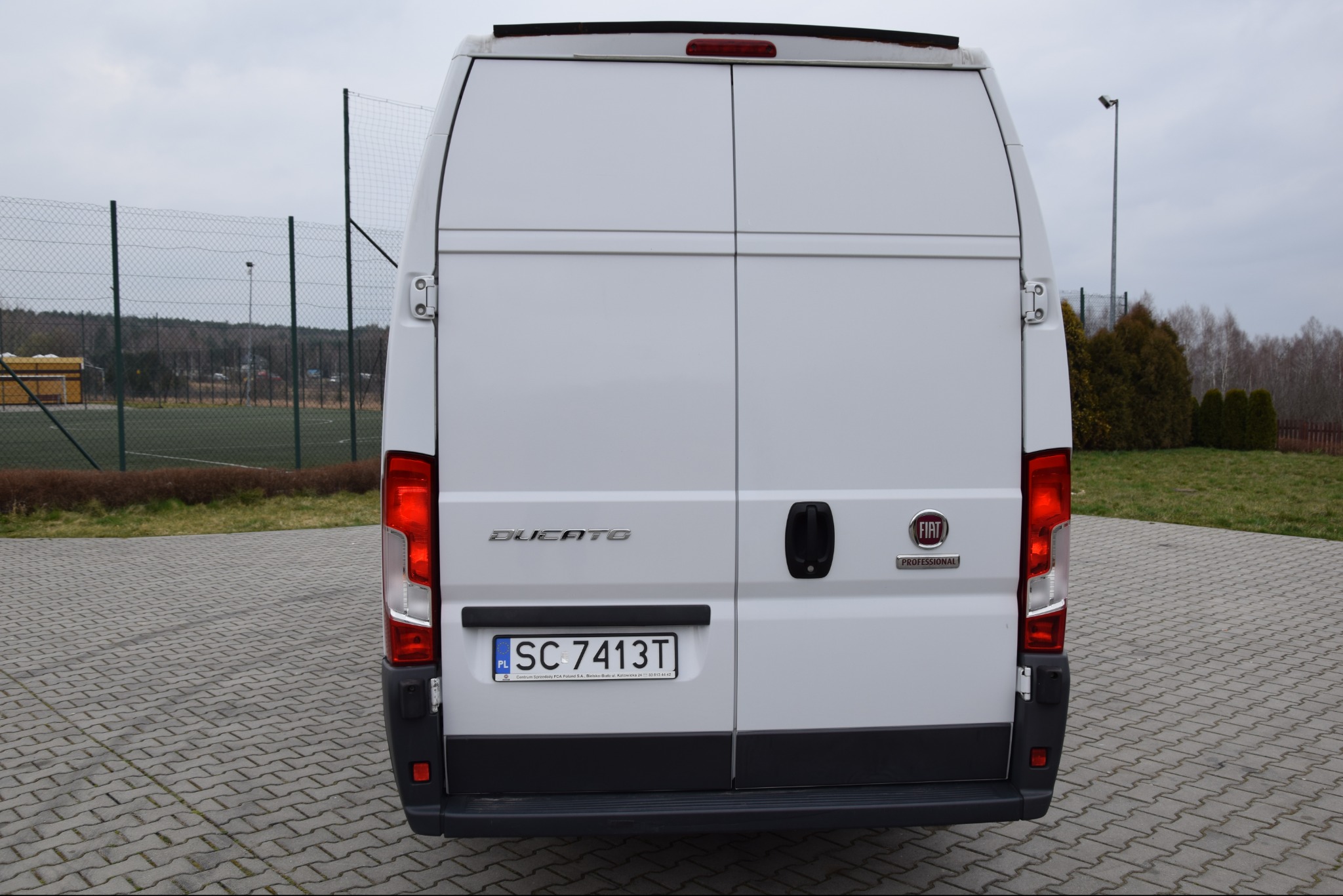 Tył białego samochodu dostawczego Fiat Ducato Professional z widoczną tablicą rejestracyjną SC 7413T na tle boiska sportowego.