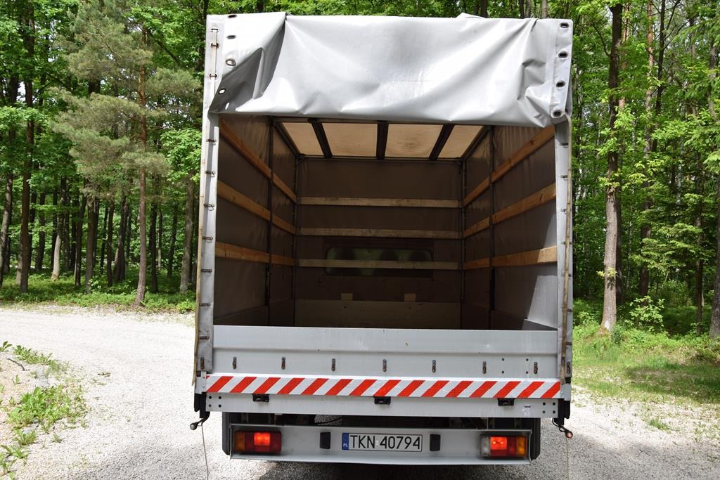 Widok z tyłu pustej przestrzeni ładunkowej samochodu ciężarowego z plandeką na tle lasu, widoczna tablica rejestracyjna TKN 40794 oraz elementy odblaskowe.