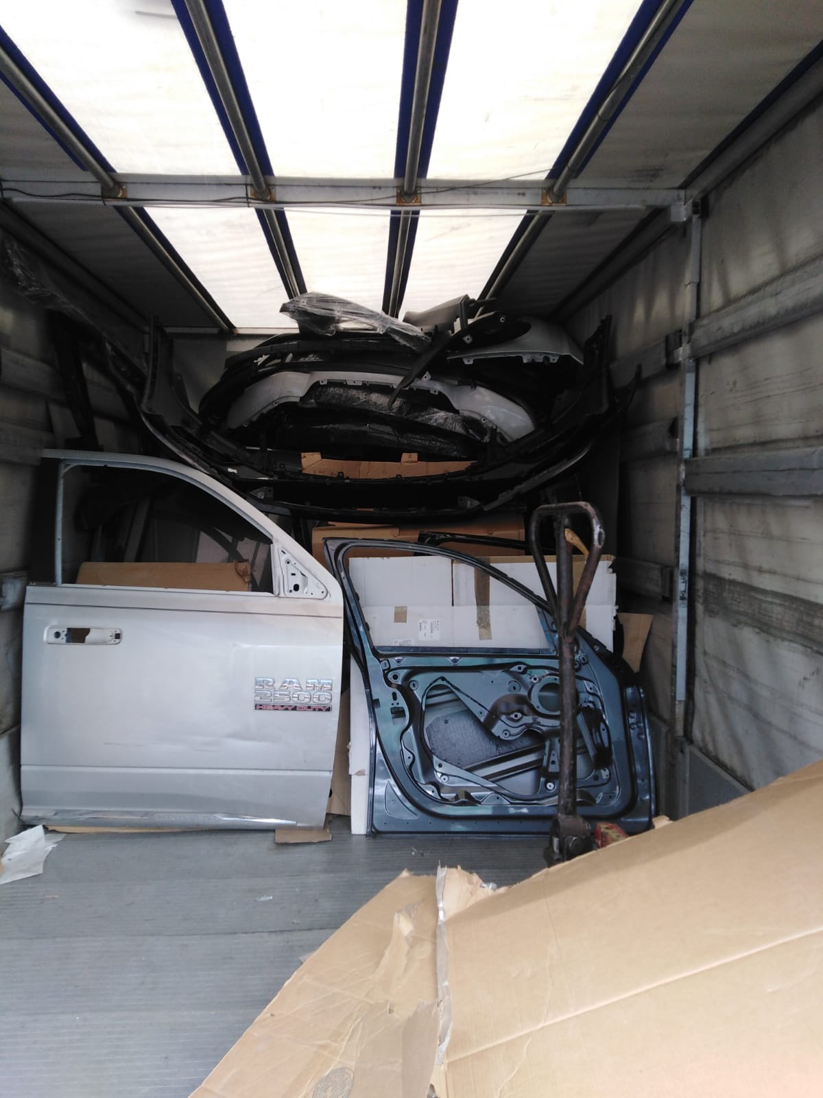 Wnętrze ciężarówki załadowane częściami samochodowymi, w tym drzwiami (srebrne z napisem RAM 2500 Heavy Duty i zielone bez tapicerki) oraz elementami karoserii, zabezpieczonymi w folii.