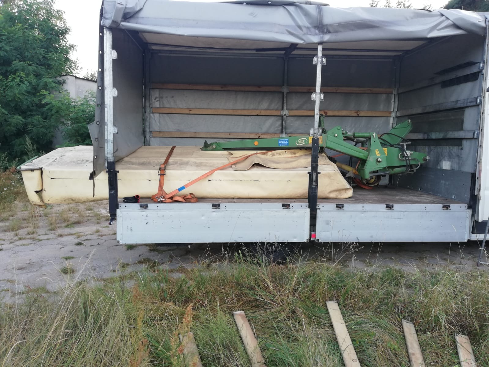 Zielona kosiarka rotacyjna marki WOLL zabezpieczona pomarańczowymi pasami transportowymi w przestrzeni ładunkowej ciężarówki z plandeką.