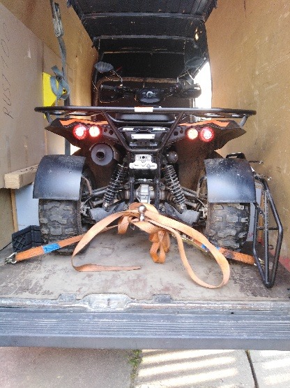 Czarny quad zabezpieczony pasami transportowymi wewnątrz drewnianej furgonetki, widok z tyłu, włączone światła stop.