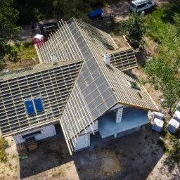 Widok z drona na budowę domu jednorodzinnego z elementami dachu w trakcie montażu, widoczne okna dachowe i kominy, teren wokół częściowo zagospodarowany, materiały budowlane składowane obok.