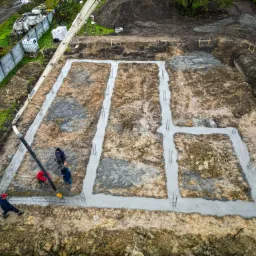 Zalewanie betonem fundamentów domu jednorodzinnego z użyciem pompy do betonu, widok z góry na wykop z ziemi i zbrojenia.