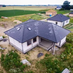 Niedokończony dom jednorodzinny z ciemnoszarą dachówką, widok z lotu ptaka, na tle pola uprawnego i innych budynków w stanie surowym otwartym, częściowo ocieplony styropianem.