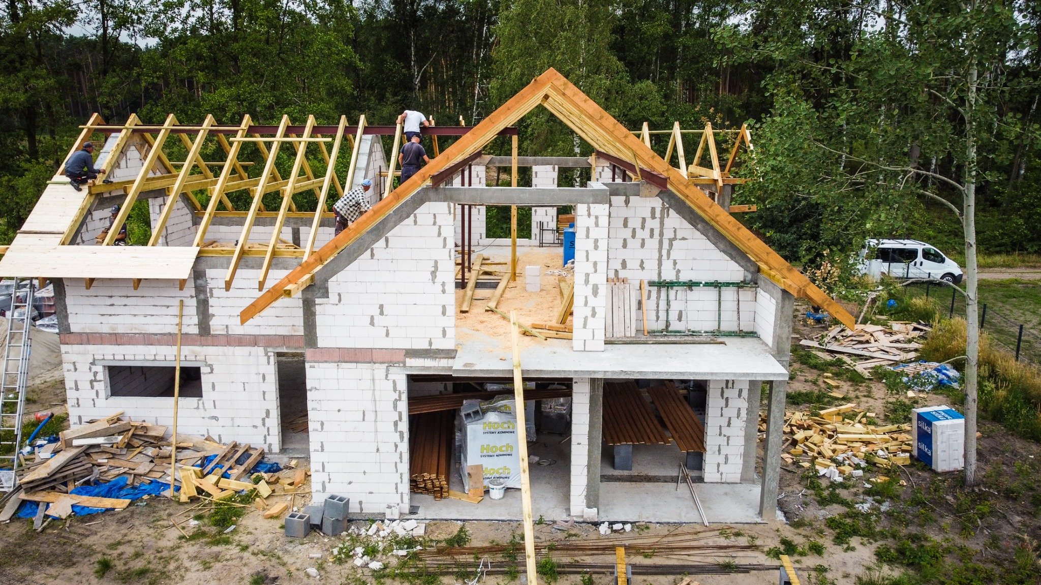 Budowa domu jednorodzinnego z białych bloczków: widok z góry na konstrukcję dachu z drewnianych belek, pracujących robotników na wysokości, częściowo wzniesione ściany parteru i poddasza, materiały...