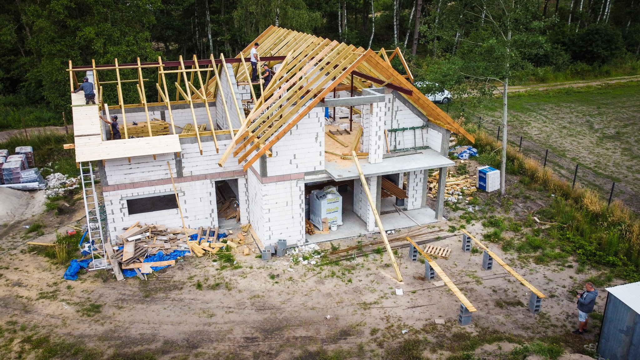 Wznoszenie domu z białych bloczków, widoczne drewniane elementy konstrukcji dachu, ekipa budowlana na dachu, teren budowy z materiałami i narzędziami, widok z lotu ptaka.