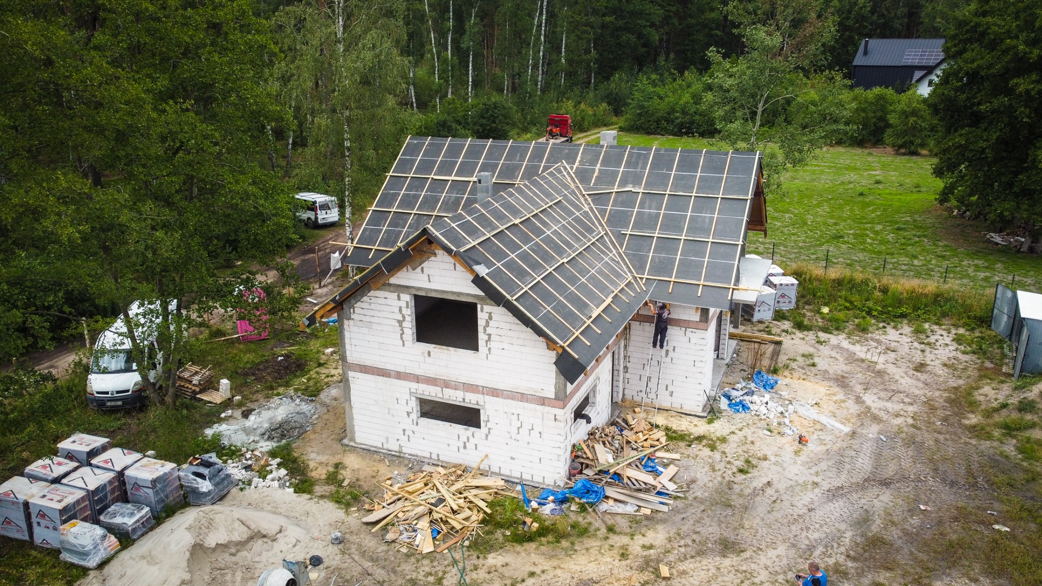 Budowa domu z białego bloczka, widok z góry, na dachu wstępne deskowanie, pracownik na drabinie, w tle las i inne budynki.