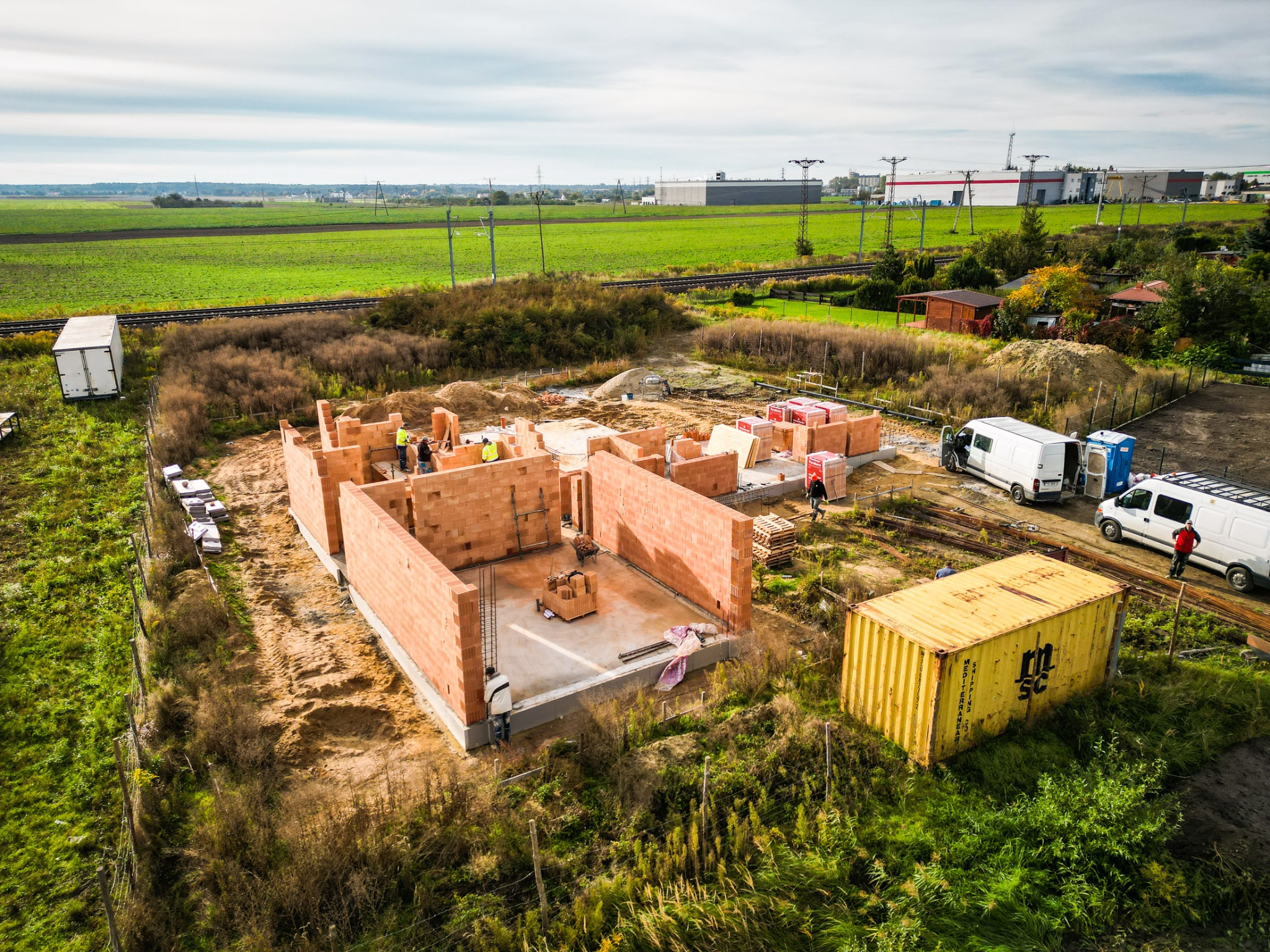 Wczesny etap budowy domu z czerwonej cegły, widok z góry, z widocznymi pracownikami w żółtych kamizelkach, materiałami budowlanymi na paletach, kontenerem magazynowym i zaparkowanymi samochodami...