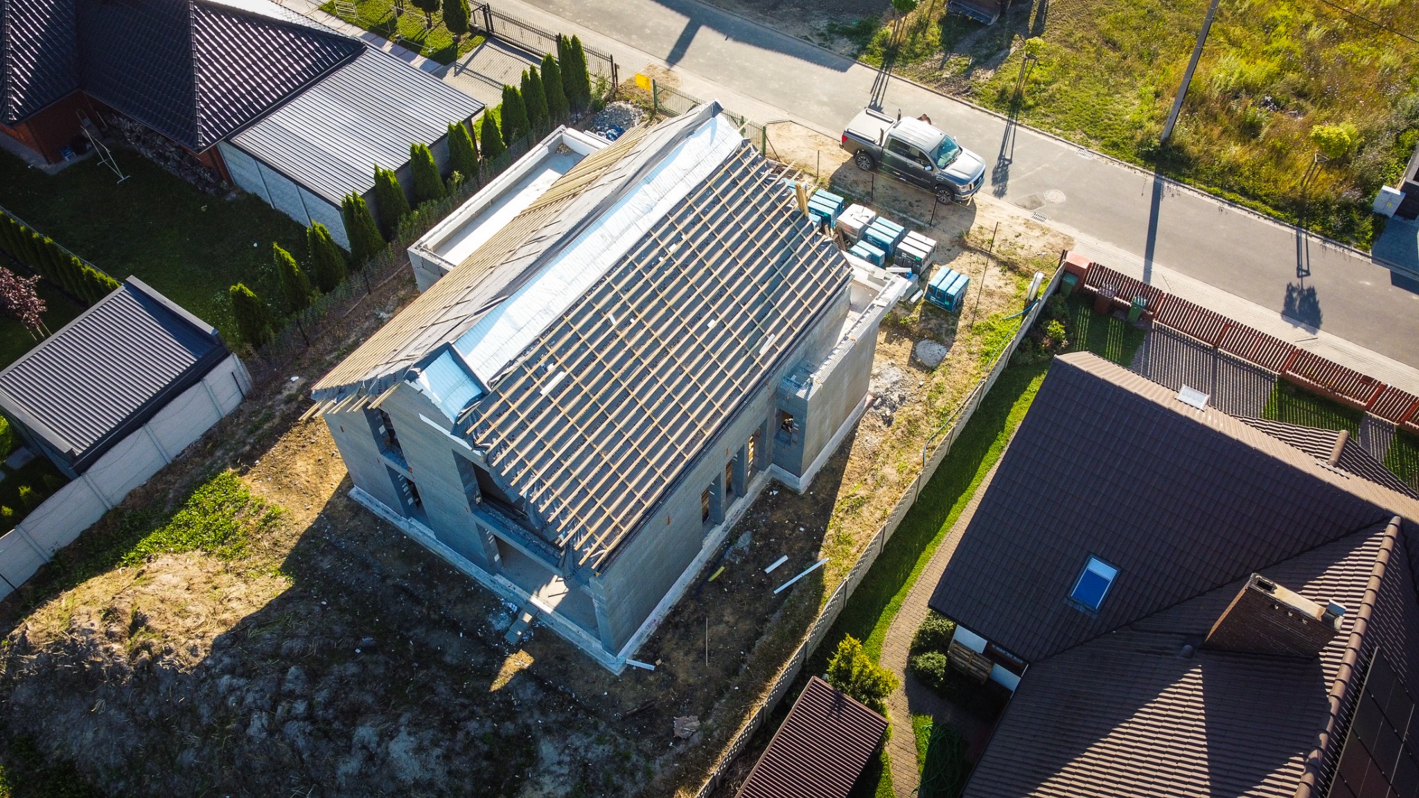 Zdjęcie z lotu ptaka przedstawia budowę domu w stanie surowym otwartym z widoczną konstrukcją dachu, pustakami i częściowo folią dachową, obok zaparkowany pickup i palety z materiałami budowlanymi,...