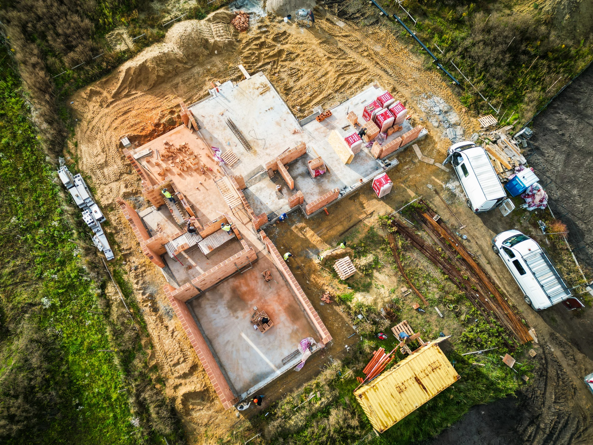 Widok z lotu ptaka na plac budowy domu z cegły, z widocznymi fundamentami, częściowo wzniesionymi ścianami, paletami z materiałami budowlanymi, pracownikami w odblaskowych kamizelkach,...