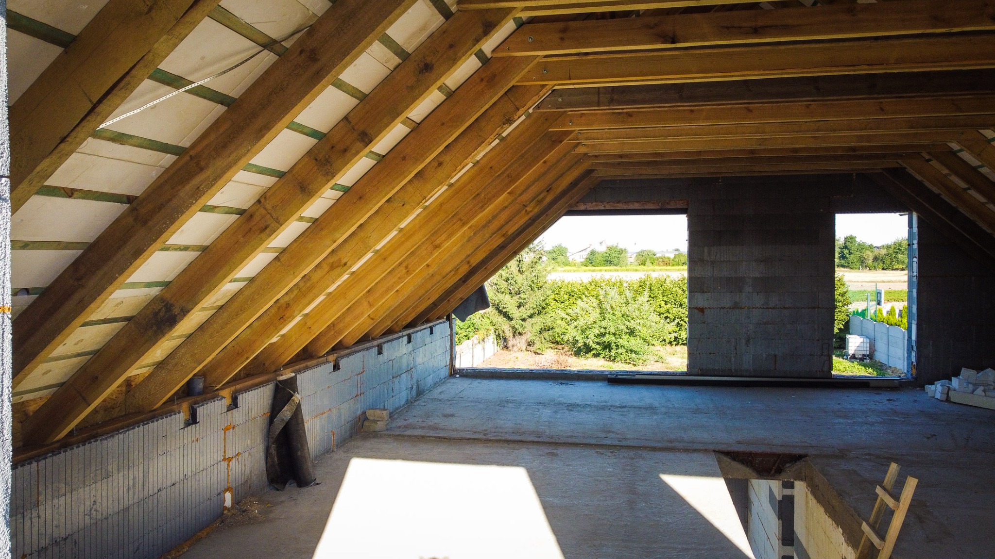 Surowy stan poddasza w nowo budowanym domu, z widoczną konstrukcją dachu z drewnianych belek i ociepleniem, ściany z bloczków betonowych, otwory okienne z widokiem na zieleń, częściowo zalana...