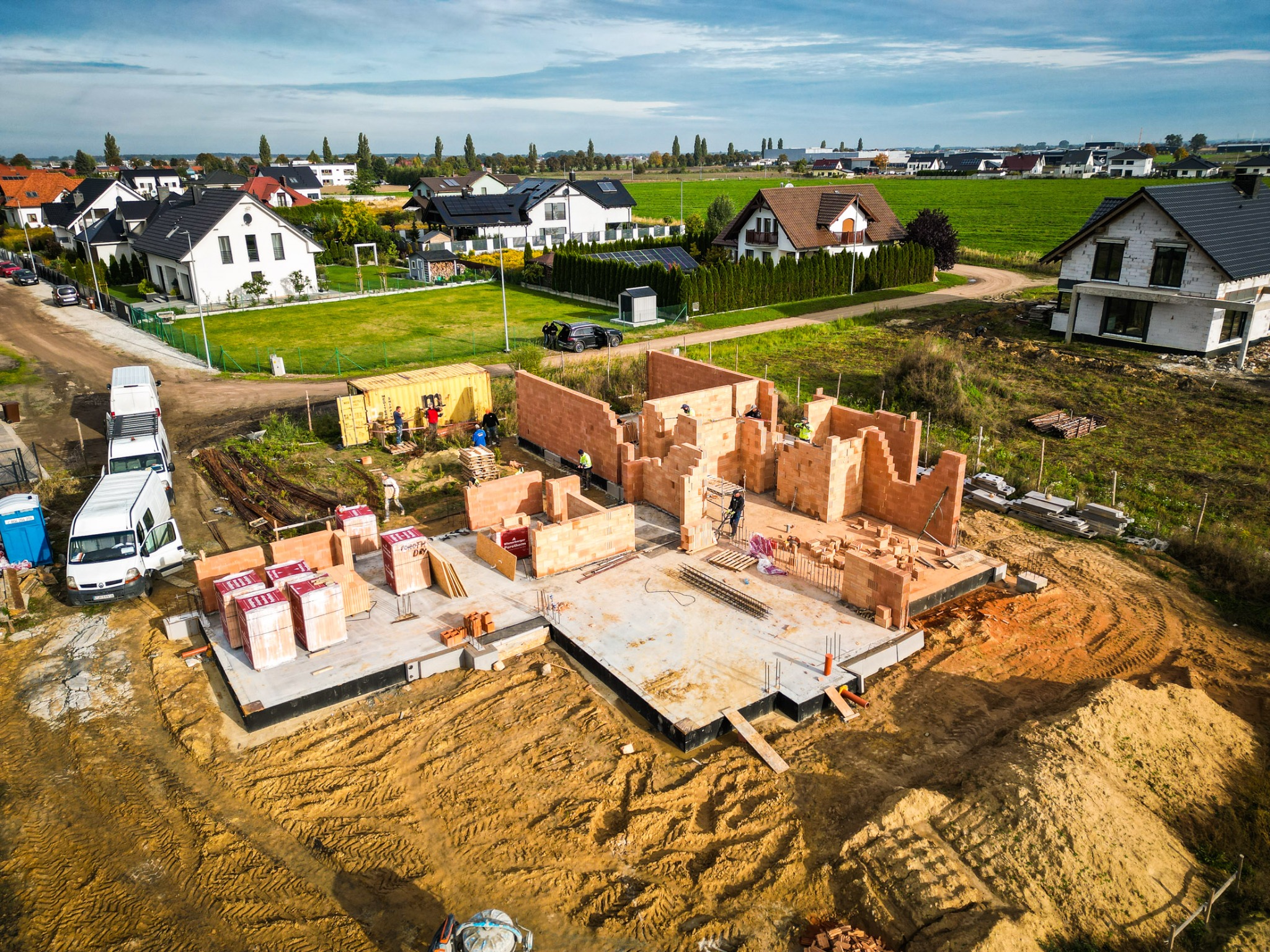 Budowa domu jednorodzinnego z czerwonej cegły, widok z lotu ptaka na fundamenty, ściany i plac budowy z materiałami, w tle zabudowa osiedlowa i zielone pola.
