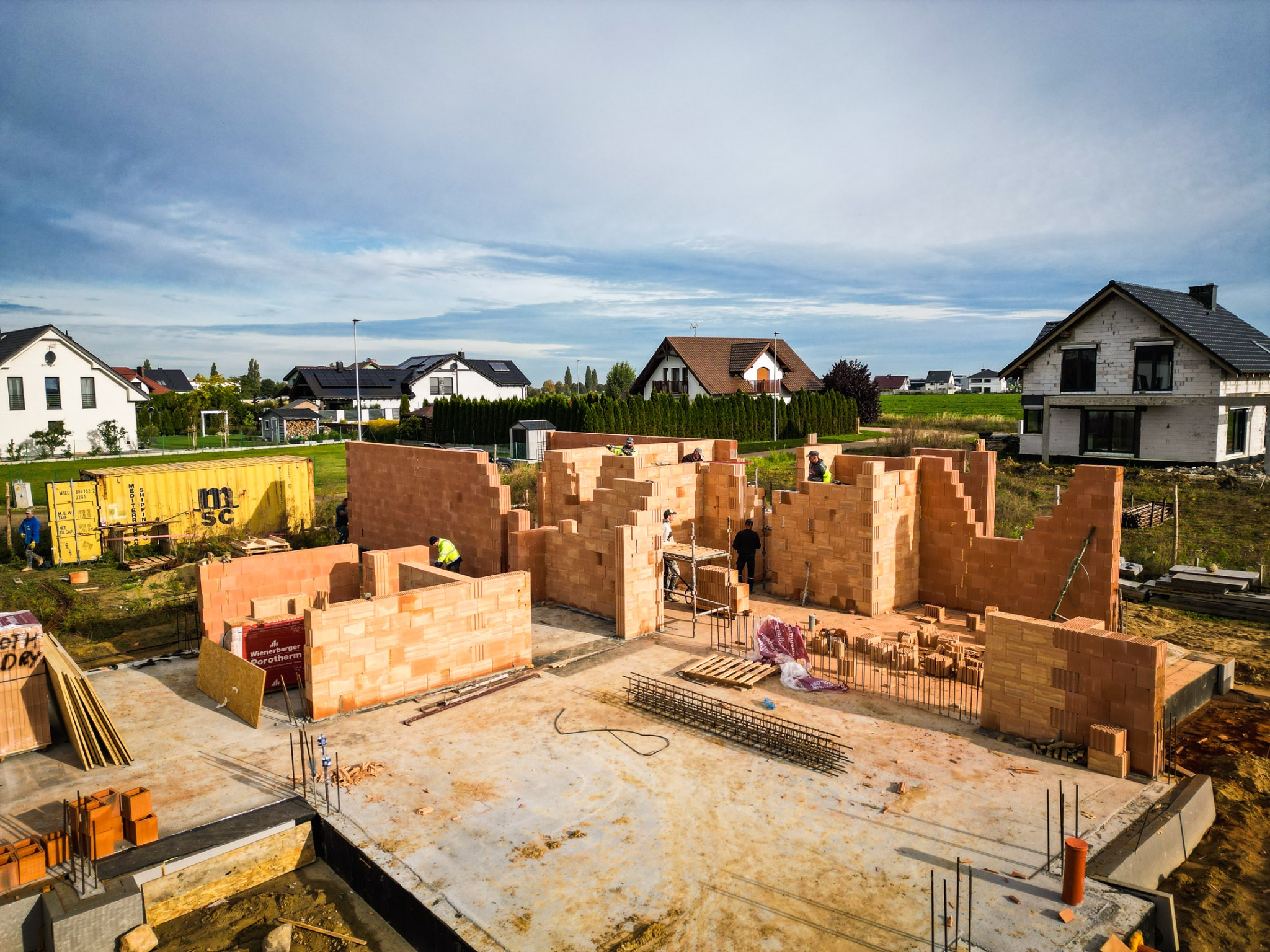 Budowa domu murowanego z czerwonej cegły Porotherm, widoczne fundamenty, ściany w trakcie wznoszenia, zbrojenia, oraz pracujący robotnicy w kamizelkach odblaskowych na tle zabudowy jednorodzinnej...