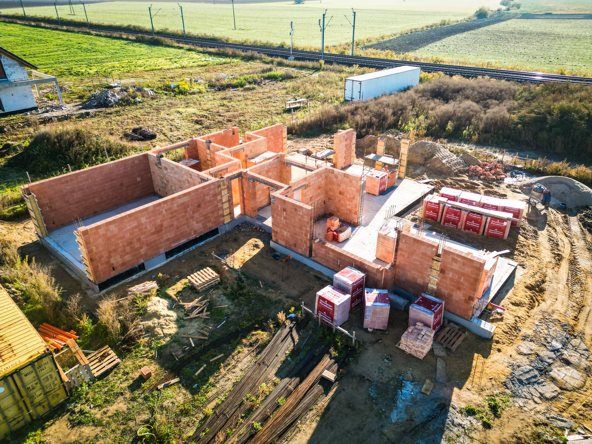 Wysoka perspektywa na budowę domu z czerwonej cegły Porotherm, widoczne fundamenty i ściany parteru, na działce leżą materiały budowlane i stoi kontener, w tle linia kolejowa i pole uprawne.