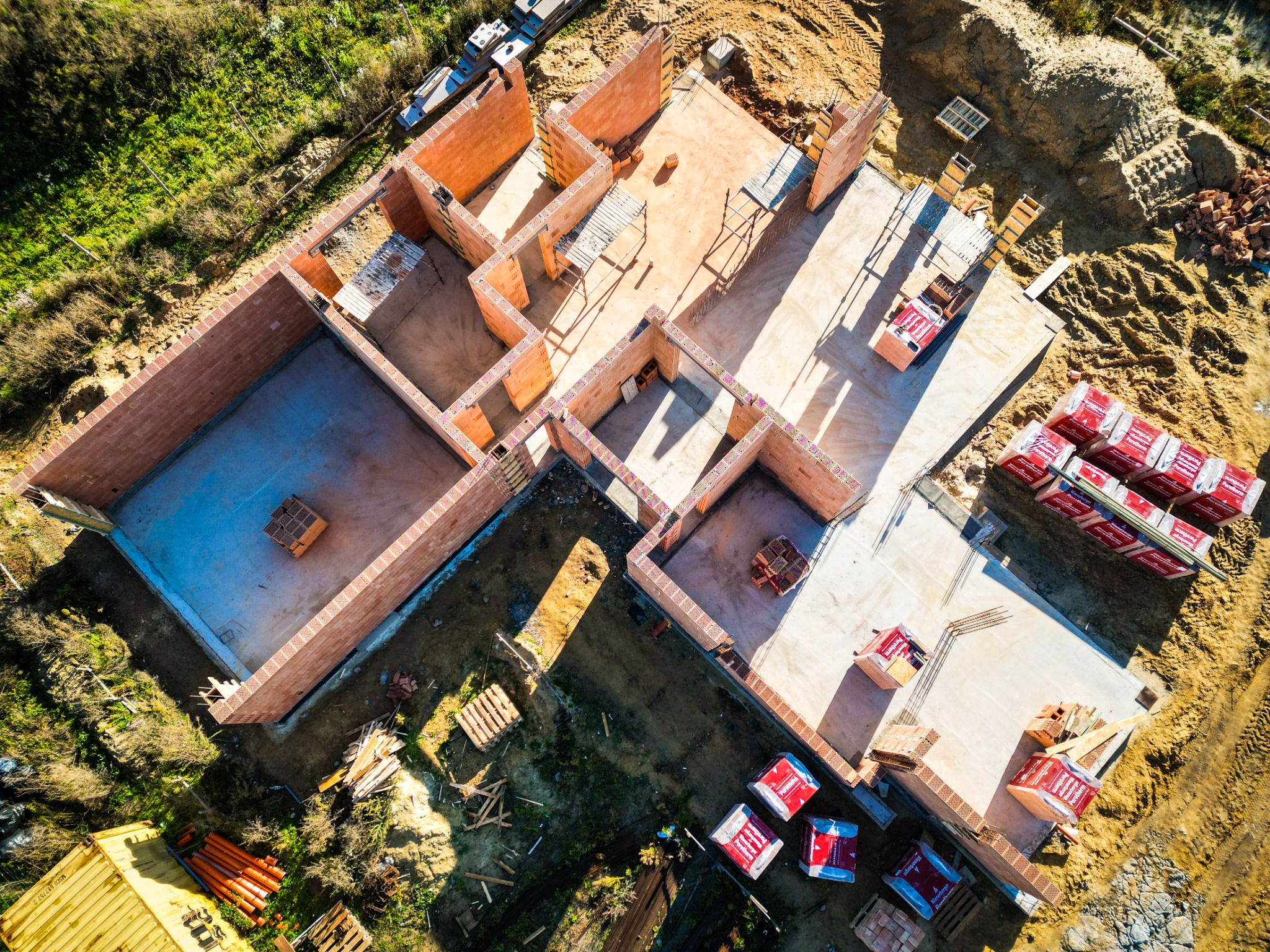 Widok z drona na budowę domu z czerwonej cegły, widoczne fundamenty i ściany parteru, palety z materiałem budowlanym, teren budowy z zielenią i piaskiem.