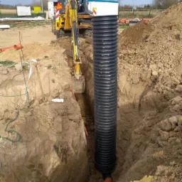 Wkopana w ziemię karbowana rura z tworzywa sztucznego, połączona z pomarańczową rurą kanalizacyjną, w tle koparka podczas prac ziemnych.