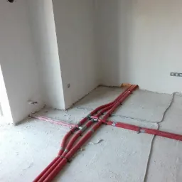 Czerwone rury instalacji grzewczej rozprowadzone po podłodze w surowym stanie w pomieszczeniu w Pieńsku, widoczne mocowania i fragmenty ścian.