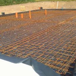 Siatka zbrojeniowa na czarnej folii, przygotowana do wylania fundamentów, z widocznymi pomarańczowymi rurami instalacyjnymi.