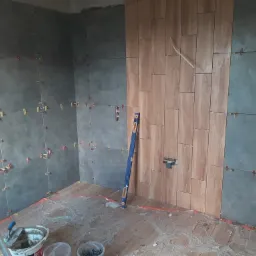 Wnętrze w trakcie prac wykończeniowych: częściowo wyłożone szarymi i drewnopodobnymi płytkami, z użyciem klipsów poziomujących; poziomica oparta o ścianę, wiadro z zaprawą i drabina w tle.