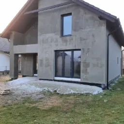 Surowy, dwukondygnacyjny dom jednorodzinny w trakcie prac elewacyjnych, z widocznymi oknami z czarnymi ramami, szarą, nieotynkowaną fasadą, brązowym dachem, częściowo stopionym śniegiem u podstawy...