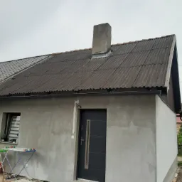 Dom z częściowo wymienionym pokryciem dachowym, nowa blacha trapezowa obok starej, falistej. Ściany zewnętrzne w stanie surowym, przygotowane do wykończenia. Widoczny komin i fragment rynny.