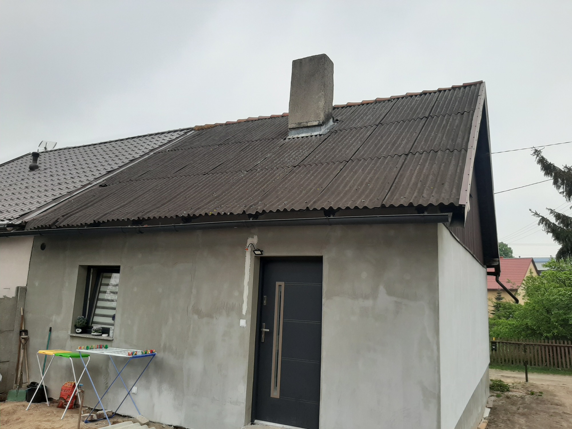 Dom z częściowo wymienionym pokryciem dachowym, nowa blacha trapezowa obok starej, falistej. Ściany zewnętrzne w stanie surowym, przygotowane do wykończenia. Widoczny komin i fragment rynny.