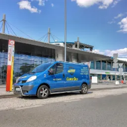 Niebieski bus Cosmo Bus z numerem telefonu na tle terminalu lotniska, z widocznym znakiem 'minibus peri>>' i fragmentem parkingu.